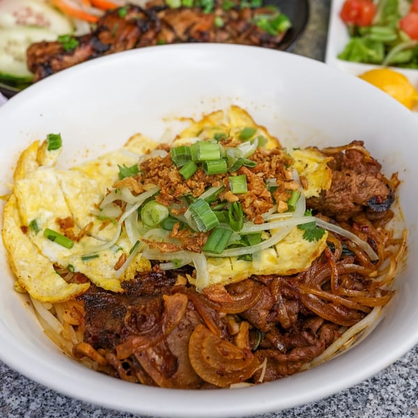 Vietnamese dish with noodles, grilled meat, fried egg, and green onions in a white bowl.