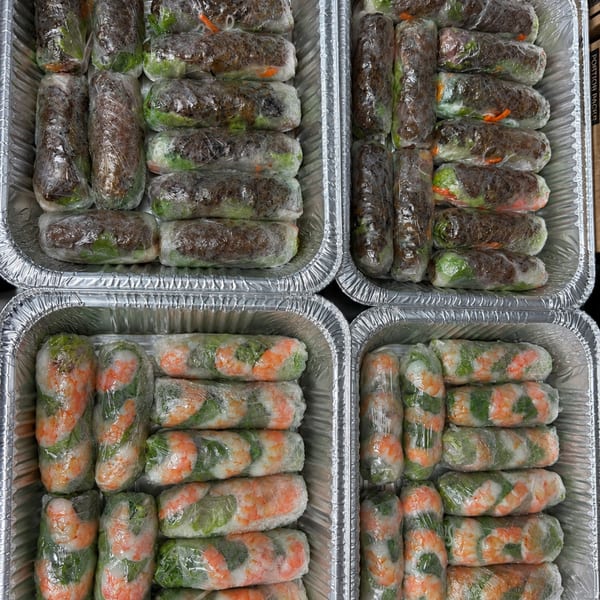 Four foil trays filled with rows of individually wrapped spring rolls, some with shrimp, others with beef.