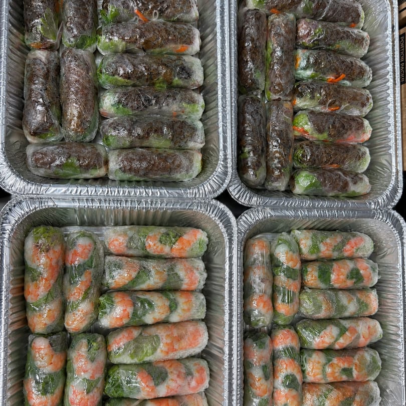 Four foil trays filled with rows of individually wrapped spring rolls, some with shrimp, others with beef.