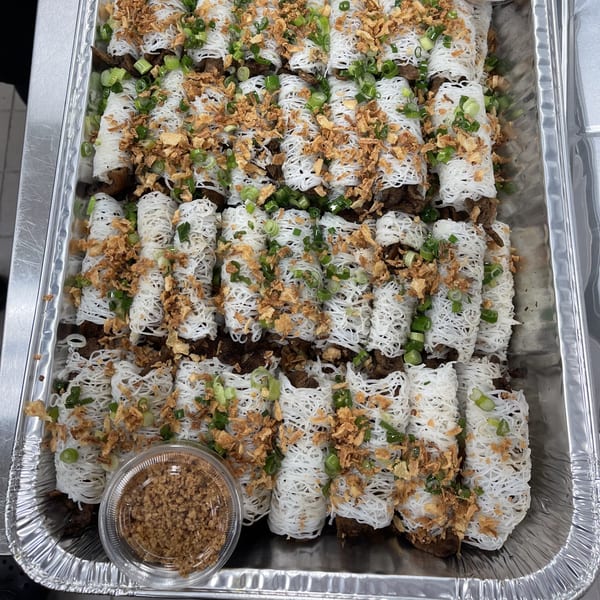 A catering tray filled with numerous noodle-wrapped rolls, garnished with green onions and crispy fried shallots.