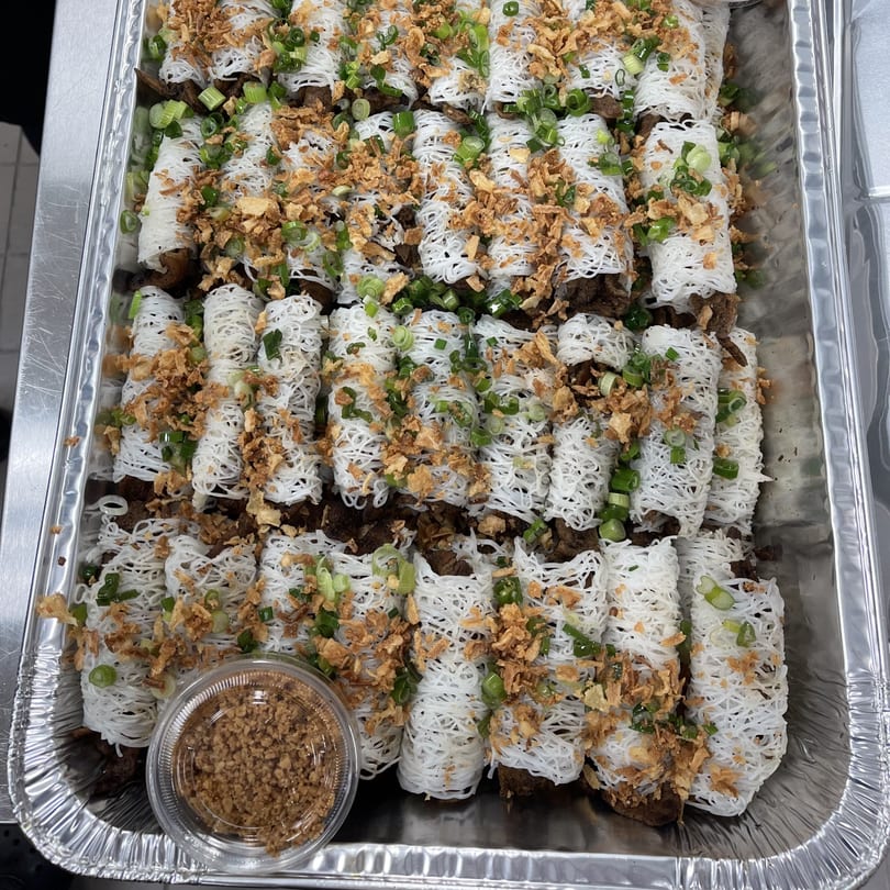 A catering tray filled with numerous noodle-wrapped rolls, garnished with green onions and crispy fried shallots.