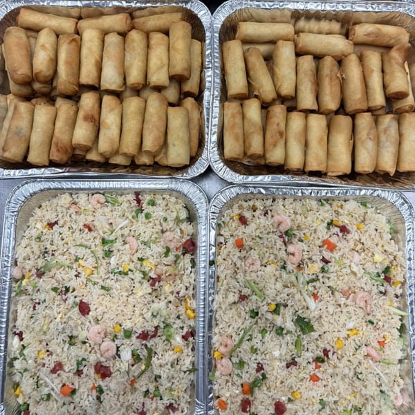 Four aluminum trays filled with golden-brown spring rolls and shrimp fried rice with vegetables.