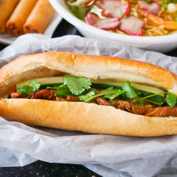 A Vietnamese banh mi sandwich with meat, cucumbers, and cilantro, with spring rolls and pho in the background.